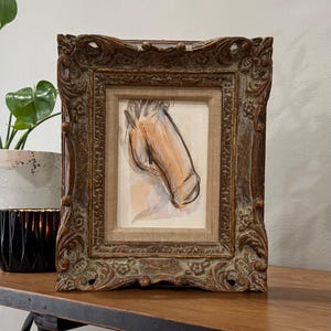 May include: An artwork in a decorative brown frame, depicting a sketch of a horse's head in brown and beige tones. The frame has an ornate design. The artwork is displayed on a wooden surface, with a plant and candle holder visible in the background.