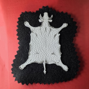 May include: A white leather patch in the shape of a cowhide rug, with a detailed illustration of a cow. The patch is set on a black felt background with a jagged edge.