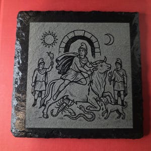 May include: Square slate coaster with a black border and a gray center. The gray center features a detailed engraving of a figure riding a bull, accompanied by other figures, a sun, a moon, and a dog.
