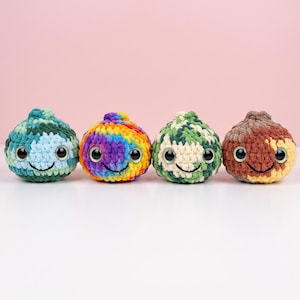 May include: Four round, crocheted plush toys arranged in a row. Each features a textured yarn surface, large black plastic safety eyes with green irises, and a stitched black smile. The colors include blue-green, rainbow, green-white, and brown-tan.
