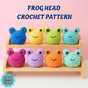 May include: Eight plush frog heads in blue, yellow, green, teal, pink, purple, orange, and mint colors arranged on a wooden shelf. The text FROG HEAD CROCHET PATTERN and LOW SEW is visible. Each frog has two dark eyes and a stitched V-shaped mouth.