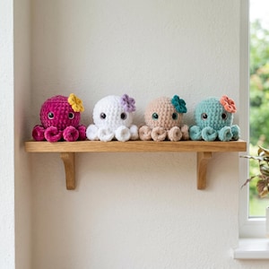 May include: Four small, round, crocheted octopus plush toys with large black eyes and curled tentacles. Each octopus features a different color: magenta with a yellow flower, white with a purple flower, beige with a teal flower, and teal with an orange flower.