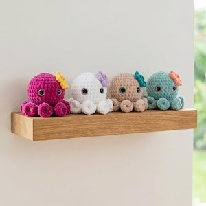 May include: Four crocheted octopus plush toys lined up in a row. Each octopus features a round head, curled tentacles, large dark eyes, and a small crocheted flower attached to the side of its head. The colors are magenta, white, tan, and teal.