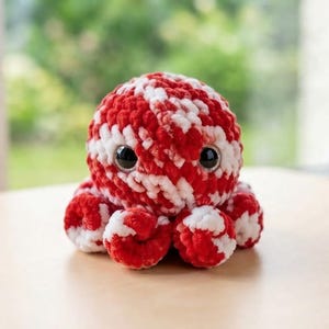 K&ouml;nnte beinhalten: Ein kleines, rundes, geh&auml;keltes Oktopus-Pl&uuml;schtier mit einem gemusterten Design aus leuchtend rotem und wei&szlig;em Garn. Das weiche, strukturierte Wesen hat zwei gro&szlig;e, gl&auml;nzende schwarze Sicherheitsaugen mit silberfarbenen R&auml;ndern an der Vorderseite.
