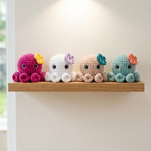 May include: Four small, crocheted octopus plush toys lined up in a row. Each octopus features a textured yarn body, large dark eyes, and a small crocheted flower attached to the side of its head. The colors are magenta, white, beige, and teal.