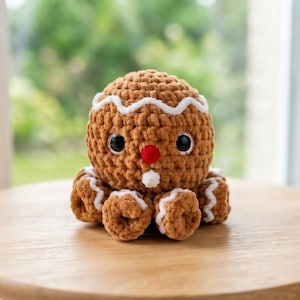 K&ouml;nnte beinhalten: Ein braunes, geh&auml;keltes Oktopus-Stofftier im Lebkuchen-Design. Es verf&uuml;gt &uuml;ber wei&szlig;e, wellenf&ouml;rmige Garnverzierungen, zwei gro&szlig;e schwarze Augen mit wei&szlig;em Rand, eine kleine rote Bommelnase und einen winzigen wei&szlig;en Bommelmund auf dem runden Kopf.