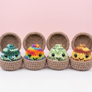 May include: Four small, round, crocheted plush creatures with large, glossy black eyes and stitched smiles, each nestled inside a matching brown crocheted shell. The creatures feature variegated yarn patterns in shades of green, rainbow, light green, and brown.