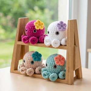 May include: Four small, round, crocheted octopus plushies with large dark eyes and a single small crocheted flower attached to the top of each head. The octopuses are magenta, white, beige, and teal, displayed on a two-tiered wooden shelf.