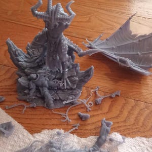 May include: A grey 3D printed resin figurine of a horned dragon-like creature standing on a rocky base, accompanied by two detached wings and several small broken fragments of resin material scattered around the central figure.