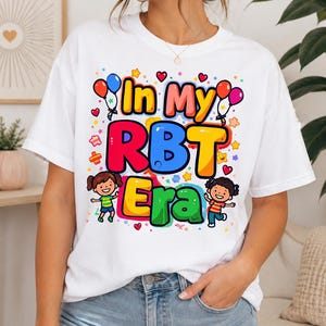 Op de afbeelding: Wit T-shirt met de tekst "In My RBT Era" in kleurrijke letters. Het ontwerp bevat cartoon illustraties van kinderen, ballonnen, sterren en harten. Een casual kledingstuk voor dagelijks gebruik.