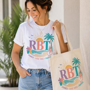 May include: A white t-shirt and a matching canvas tote bag, both featuring a colourful graphic with the text RBT, a sun, a palm tree, a puzzle piece, and a heart. The graphic includes the words SUMMER, GOOD VIBES, and PURPOSE in a circular arc.