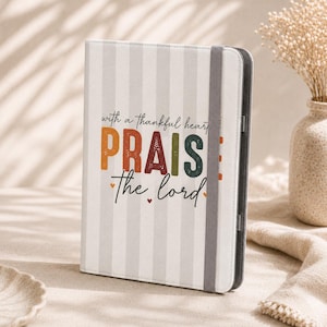 May include: A vertical notebook with a light gray and white striped cover. The front features the text "with a thankful heart PRAISE the lord" in orange, red, and dark green, accented by small hearts. A gray elastic band secures the notebook closed.