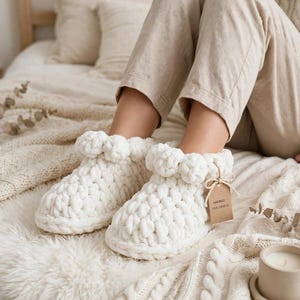 Op de afbeelding: Een paar off-white, grof gebreide chenille pantoffels met een dikke, textuurrijke boord rond de enkel. Een klein, rechthoekig bruin papieren label is met een touwtje bevestigd en bevat de tekst "HANDMADE 100% CHENILLE".