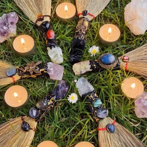 May include: An overhead shot of a collection of decorative brooms with ornate handles, surrounded by lit candles, crystals, and flowers on a bed of green grass. The brooms have natural straw bristles and are adorned with various gemstones and beads.