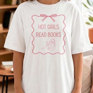 Puede incluir: Camiseta blanca con un gráfico rosa que dice "HOT GIRLS READ BOOKS". El gráfico presenta un lazo rosa y un dibujo de una persona leyendo. La camiseta tiene un corte holgado y de gran tamaño.