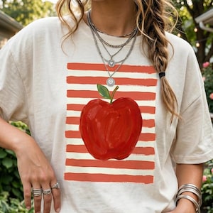 Puede incluir: Camiseta color crema con un gran gráfico de manzana roja con una hoja verde, sobre un fondo de rayas horizontales rojas y blancas. La camiseta tiene un corte holgado.