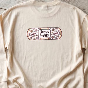 Puede incluir: Camiseta de manga larga color crema con un diseño de vendaje con las palabras "Jesus heals". El vendaje está decorado con motivos florales y de corazón, junto con las palabras "Faith" y "Hope".