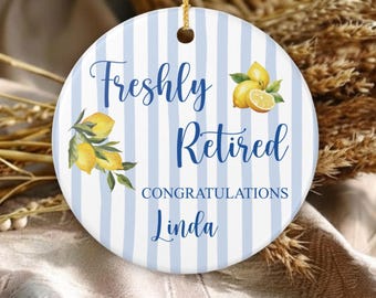 May include: A round white ceramic ornament featuring light blue vertical stripes and yellow lemon illustrations with green leaves. The text reads Freshly Retired CONGRATULATIONS Linda in blue script and block lettering. It hangs by a gold cord.