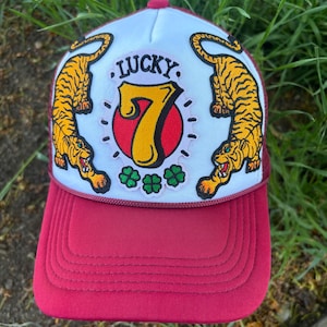 Op de afbeelding: Een bordeauxrode en witte truckerpet met een "Lucky 7" patch. De patch heeft een gouden nummer 7, twee oranje tijgers en het woord "LUCKY". De pet heeft ook drie groene klavers.
