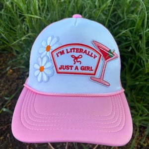 Op de afbeelding: Roze en witte truckerpet met een roze klep. De pet heeft de tekst "I'M LITERALLY JUST A GIRL" in het rood, samen met cocktail- en madeliefjesaccenten. De pet heeft een roze knoop aan de bovenkant en een wit touwdetail.