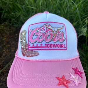 May include: Pink and white trucker hat with a pink brim and a patch featuring the word "Coors" in pink script, a bull skull, and the word "Cowgirl". A rhinestone boot and pink stars adorn the hat.
