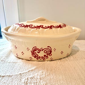 May include: An oval ceramic casserole dish with a matching lid, featuring a cream-colored base and red painted decorative patterns. The design includes a central heart motif, small dot accents, and a scalloped rim detail on the lid and body.