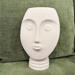 May include: A light beige, textured ceramic vase shaped like a human face. The vase features a smooth, oval-shaped head with recessed circular eyes, a long straight nose, and small lips. The base of the vase is cylindrical with two horizontal rings.