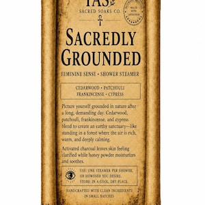 May include: A rectangular product label with a tan, parchment-like scroll design. The text reads TAS SACRED SOAKS CO. SACREDLY GROUNDED FEMININE SENSE SHOWER STEAMER. It lists cedarwood, patchouli, frankincense, and cypress, plus usage instructions.