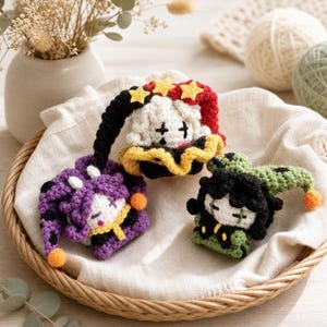 May include: Three small crocheted dolls with jester-style hats. One doll has a purple and black hat with orange pom-poms, one has a red and black hat with yellow stars, and one has a green and black hat with orange pom-poms, all with stitched faces.