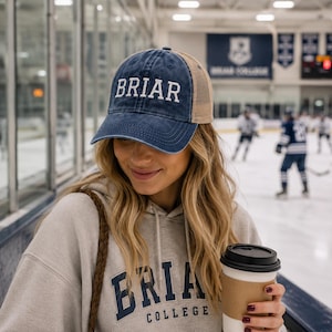 May include: A navy blue baseball cap with a tan mesh back and the word BRIAR embroidered in white on the front. The cap features a distressed finish on the brim and crown, with a structured front panel and an adjustable strap at the back.