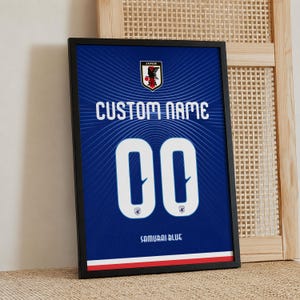 May include: A framed rectangular poster featuring a blue background with a radiating line pattern. It displays the Japan Football Association logo, the text CUSTOM NAME, the number 00, the text SAMURAI BLUE, and a red and white stripe at the bottom.