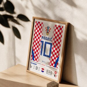 May include: A framed poster featuring a red and white checkered pattern inspired by the Croatian national team jersey. The design includes the name MODRIĆ, the number 10, the HNS crest, and player statistics for Luka Modrić with a small soccer field icon.