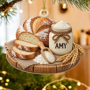 Può includere: Una decorazione natalizia rotonda in ceramica con una pagnotta di pane a fette, un vasetto etichettato AMY pieno di farina bianca, un cucchiaio di legno e spighe di grano su un tagliere di legno. L'ornamento pende da un cordino dorato.