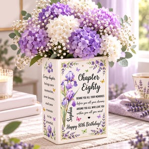 May include: A rectangular white ceramic vase filled with purple and white hydrangea and baby's breath flowers. The vase features printed iris flowers, butterflies, and text that reads: Chapter Eighty, Sophia, and a quote about memories and dreams.