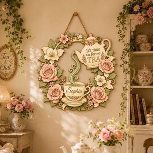 May include: A circular wall hanging featuring a floral wreath with pink roses and green leaves, surrounding a teapot pouring liquid into a teacup. The teapot has the text 'It's time for TEA' and the teacup is personalised with the name 'Sophia'.