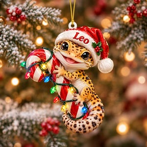 May include: A hanging Christmas ornament featuring a spotted leopard gecko wearing a red and white Santa hat with the name Leo printed on the brim. The gecko is holding a red and white candy cane wrapped in a string of small, colourful LED lights.