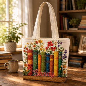 May include: A cream-coloured quilted tote bag featuring a printed design of colourful book spines arranged in a row with vibrant wildflowers growing from the top. The name Sandy is written in red cursive script across the bottom of the books.