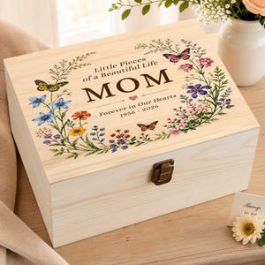 Puede incluir: Una caja de recuerdos rectangular de madera clara con tapa abatible y cierre de metal. La tapa presenta un diseño de corona floral con mariposas y el texto: Little Pieces of a Beautiful Life, MOM, Forever in Our Hearts, 1956-2026.