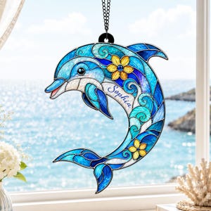 Peut inclure: Un ornement en forme de dauphin style vitrail aux nuances de bleu et de turquoise avec deux fleurs jaunes. Le nom Sophia est inscrit en lettres cursives sur le flanc du dauphin. Il est suspendu par une chaîne noire devant une fenêtre.