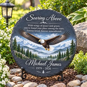 May include: A round black slate memorial plaque featuring a bald eagle in flight over a mountain forest. The text reads: Soaring Above, With wings of peace and grace, you've found your place among the stars. Forever loved, forever remembered. Michael James 1979-2026.