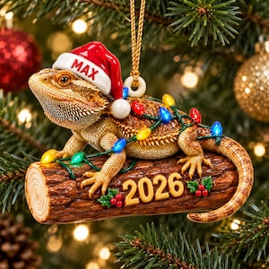May include: A bearded dragon Christmas ornament featuring the lizard wearing a red and white Santa hat with the name MAX printed on it. The lizard rests on a brown log labelled 2026, decorated with green holly leaves, red berries, and colourful lights.