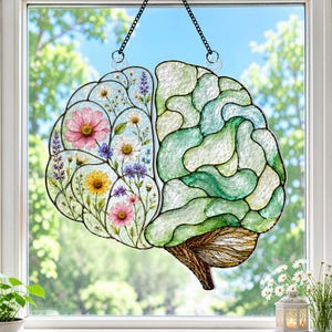 May include: A stained glass suncatcher shaped like a human brain. The left hemisphere features a clear background with colourful floral illustrations of pink, yellow, and white flowers. The right hemisphere consists of textured green glass pieces.