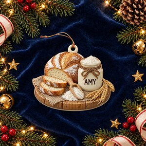 Op de afbeelding: Een ronde keramische kerstornament met een brood, een klein potje met het label AMY gevuld met witte bloem, een houten schepje en tarwearen op een houten plank. Het ornament heeft een gouden ophangkoord aan de bovenzijde.