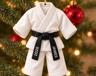 Op de afbeelding: Een wit keramieken vechtsportuniform ornament met een zwarte riem om de taille. De riem is gepersonaliseerd met de tekst 2026 aan de linkerkant en de naam JAMES aan de rechterkant. Aan de bovenkant is een gouden koordje bevestigd.