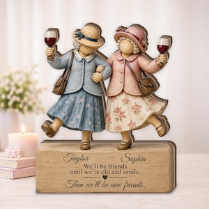 May include: A decorative figurine of two figures wearing hats, jackets, and skirts, each holding a wine glass. The base features the names Taylor and Sophia with the text: We'll be friends until we're old and senile. Then we'll be new friends.