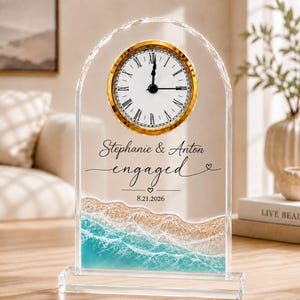May include: An arched clear acrylic clock featuring a gold-rimmed circular face with Roman numerals. The text reads Stephanie & Anton engaged 8.21.2026. The lower portion displays a printed graphic of turquoise ocean waves hitting a sandy beach.