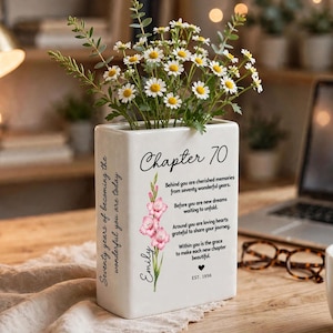 Puede incluir: Un jarrón rectangular de cerámica blanca con el texto Chapter 70, una ilustración floral rosa y un poema. El jarrón está lleno de flores blancas tipo margarita y follaje verde. El lateral del jarrón muestra el texto Seventy years of becoming the wonderful you are today.