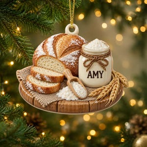 Op de afbeelding: Een hangend ornament met een ronde houten plank met een brood, twee sneetjes brood, een klein houten schepje met bloem en een cr&egrave;mekleurige keramische pot vastgebonden met touw en voorzien van de naam AMY in zwarte letters.