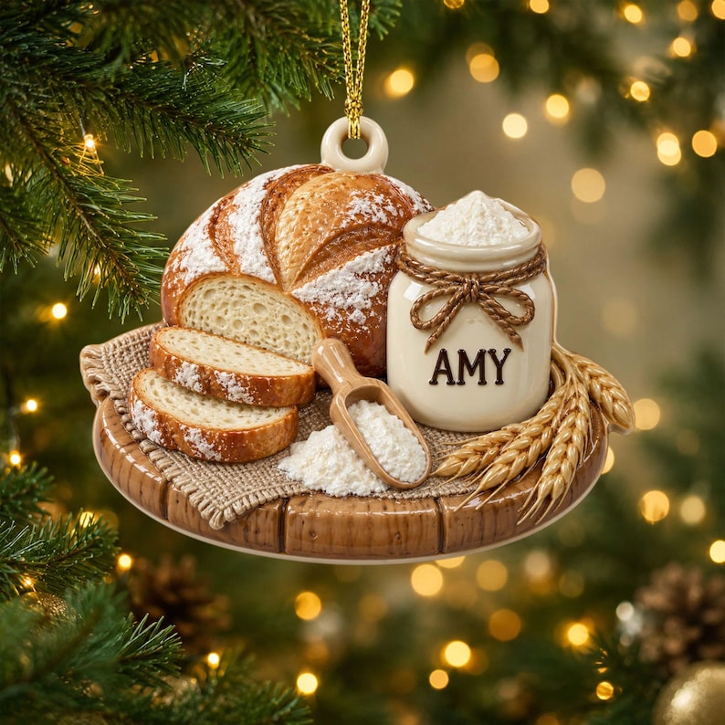 Op de afbeelding: Een hangend ornament met een ronde houten plank met een brood, twee sneetjes brood, een klein houten schepje met bloem en een cr&egrave;mekleurige keramische pot vastgebonden met touw en voorzien van de naam AMY in zwarte letters.