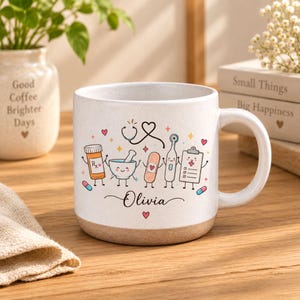 May include: A white ceramic mug with a speckled finish and a tan base, featuring a colourful graphic of cartoon medical items including a pill bottle, mortar and pestle, bandage, thermometer, and clipboard, with the name Olivia written in script.